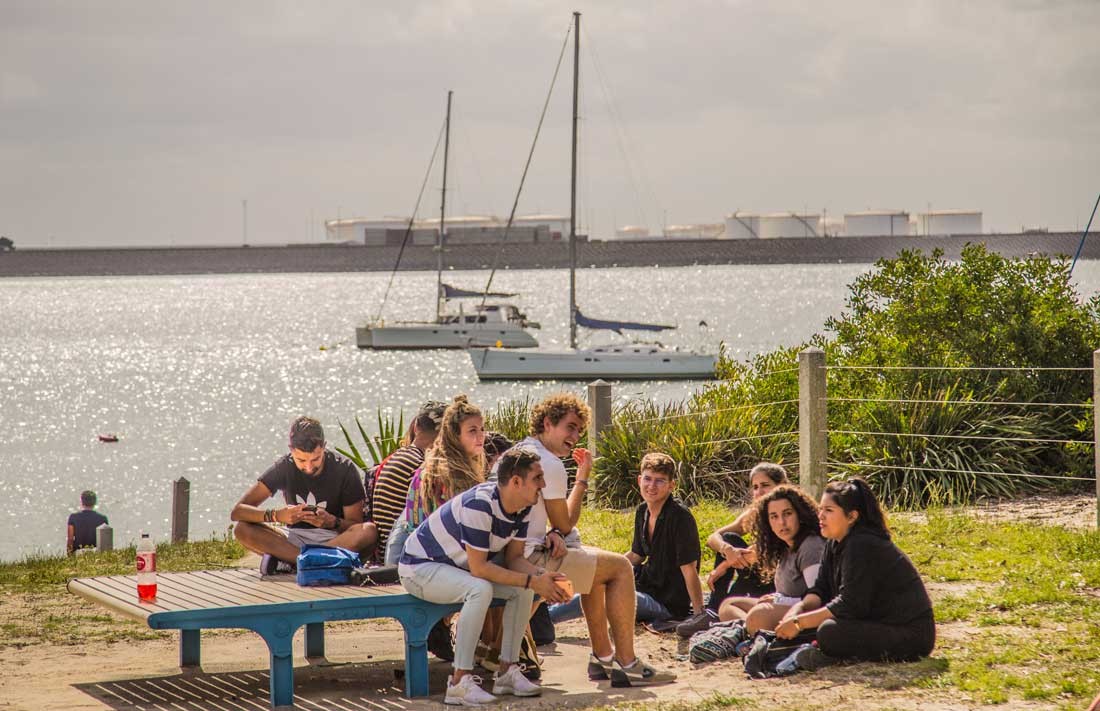 grupo de jóvenes estudiantes de tests de inglés sentados en una mesa de picnic hablando y con el mar de fondo